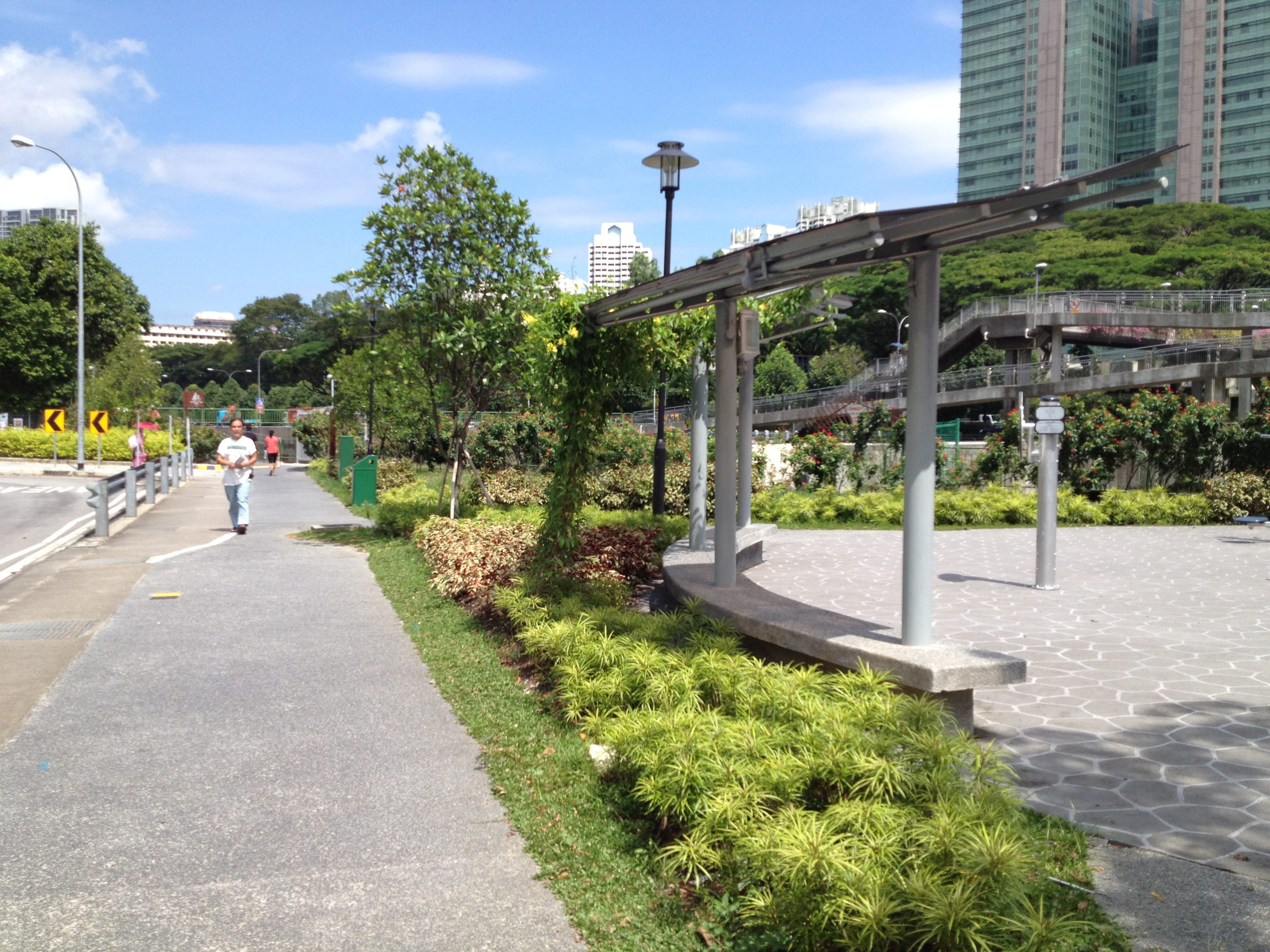 A pedestrian walks on a pathway lined with trees and plants; buildings and sky are visible in the background.